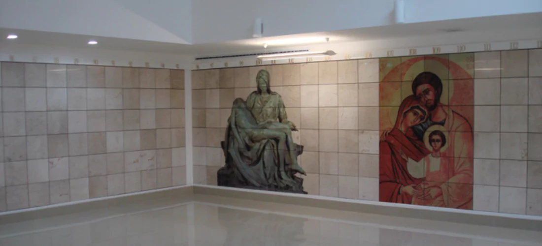 Criptas Muro de nichos en mármol beige con dos imágenes religiosas: a la izquierda, una representación de La Piedad; a la derecha, un ícono de la Sagrada Familia