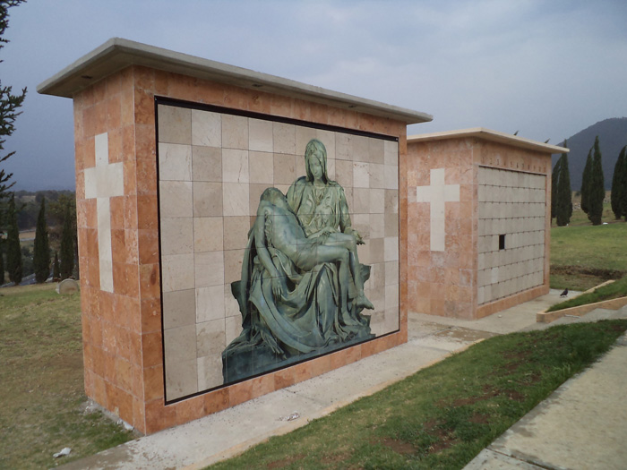 Criptas Pared de nichos exteriores en mármol con imagen de la Pietà y cruces blancas en los laterales.