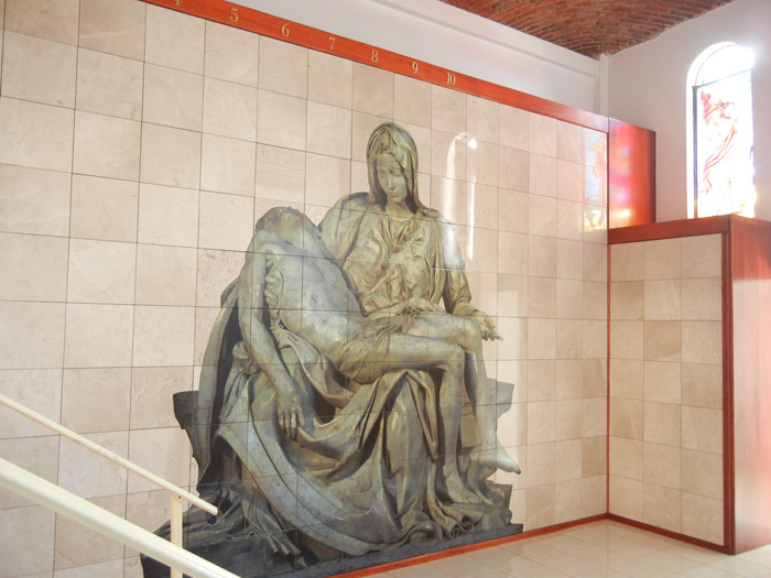Criptas Muro de mármol con imagen de la Piedad, mostrando a la Virgen María sosteniendo a Jesús, acompañado de detalles arquitectónicos que resaltan la obra en un ambiente solemne y luminoso.