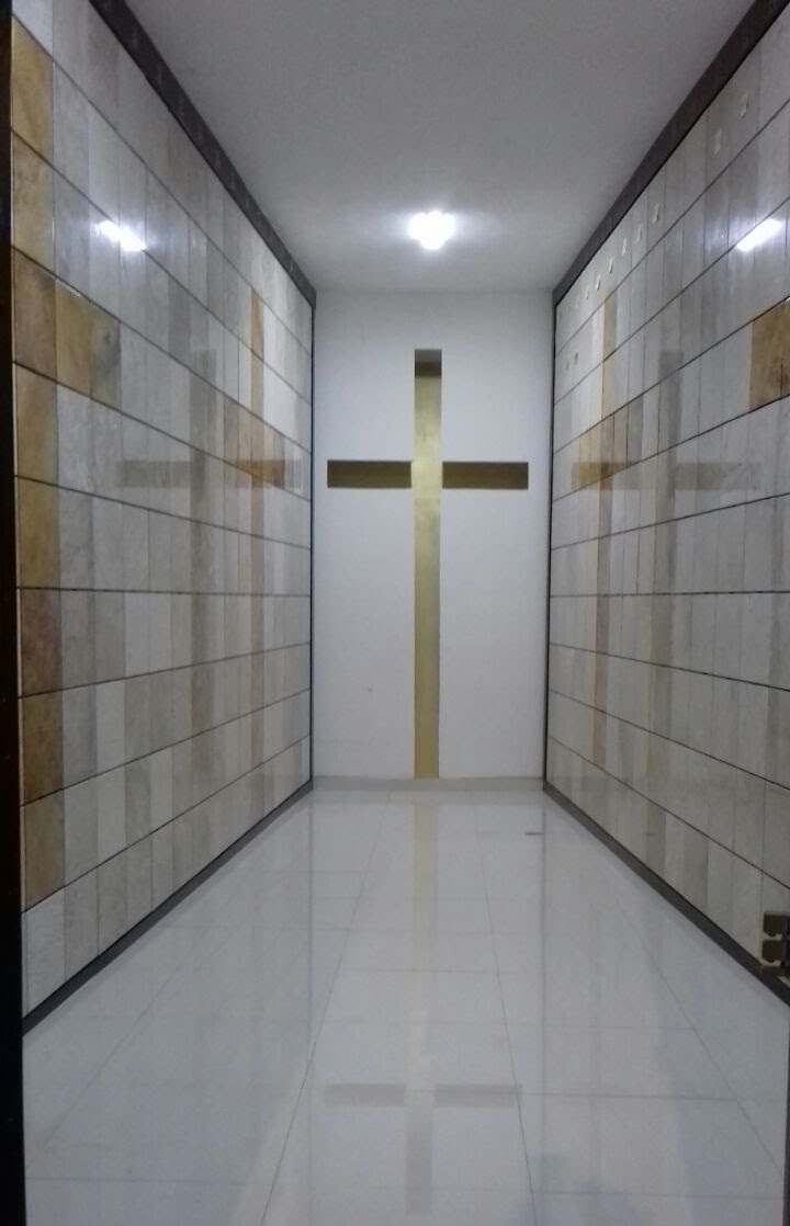 Criptas Pasillo estrecho con paredes de nichos en mármol claro, en cuyo centro se alinea una cruz dorada que destaca en el fondo, reflejada en el piso brillante