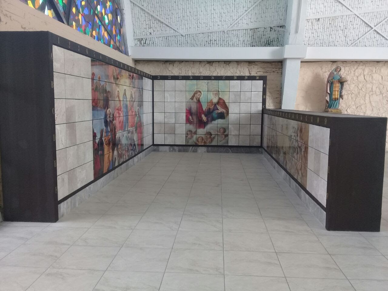 Criptas Espacio con nichos de mármol adornados con murales religiosos y una estatua al fondo, bajo una estructura de techo alto con vitrales