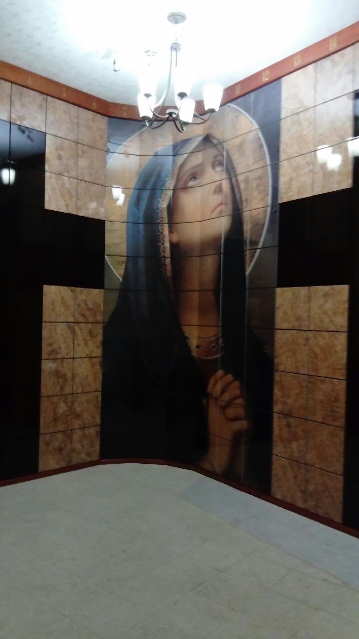Criptas Mural en mosaico de la Virgen María en actitud de oración, en una pared curva con detalles de mármol y un candelabro en el techo