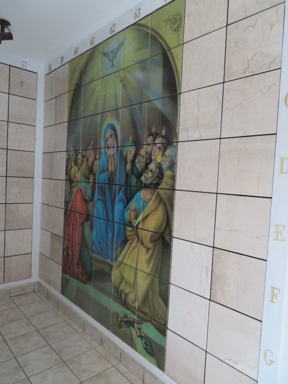 Criptas Mural en mosaico que representa a la Virgen y apóstoles en oración con una paloma en la parte superior, rodeado de baldosas lisas y numeración en los bordes