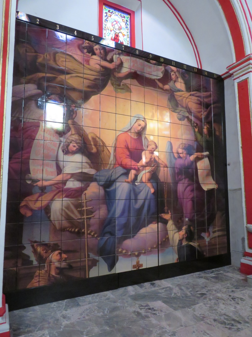 Criptas Mural en mosaico que representa a la Virgen con el Niño rodeados de ángeles, ubicado en un recinto con detalles arquitectónicos rojos y un vitral en la parte superior
